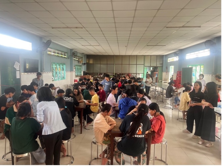 A large group of students, clustered around tables in an expansive room with windows on the right side, works collaboratively upon an engaging project.