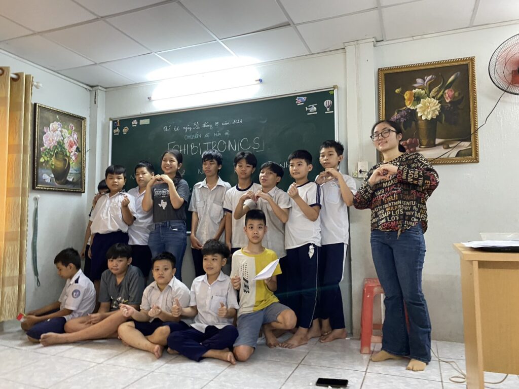 A group of children with their teacher poses in front of a green slate chalkboard with the word "Chibitronics" written in white chalk.