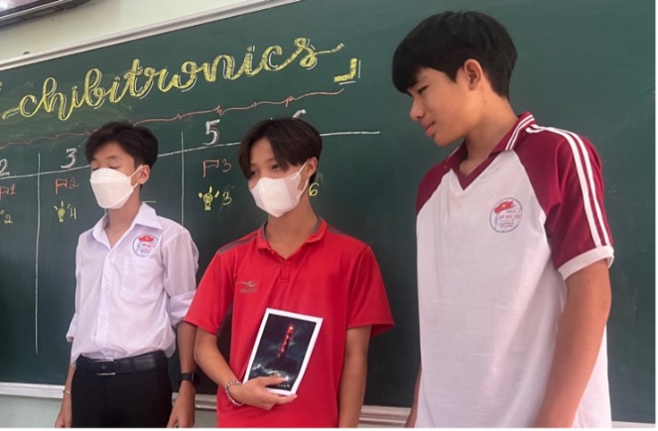 Three young adult men stand in front of a green slate chalkboard.  The one in the center holds their winning project, a glowing lighthouse with three stacked windows that glow in red.