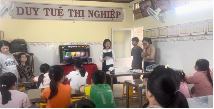 Three young adults stand in front of  classroom.  A sign on the wall in the center of the photo reads, "Duy Tue Thi Nghiep"