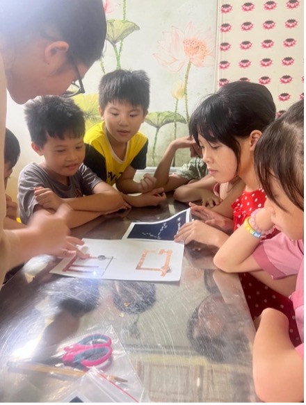 Several students gather around a piece of paper that has a circuit constructed upon it, using copper tape and a coin cell battery.