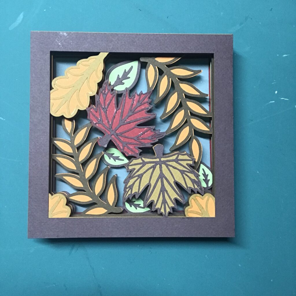A leafy, layered shadow box insert featuring differently shaped leaves in red, orange, yellow, brown, and green